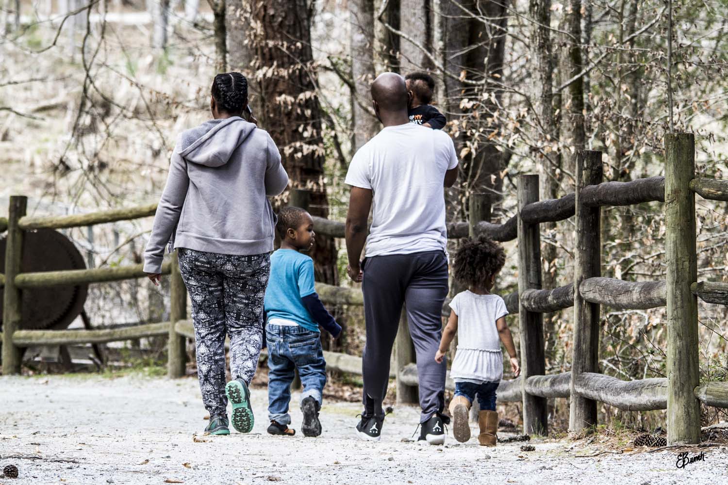 A family walks down a gravel walkway toward the wetland. It's winter and the trees are mostly bare. There is a wooden fence to their right. The family consists of a woman, a man, and three young children. They're all facing toward the wetland except one boy who holds his mother's hand and looks to the right.